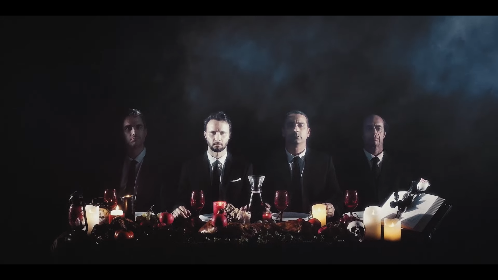 Sob fundo preto, quatro homens vestidos de fato estão sentados atrás de uma mesa repleta de alimentos e vinho, incluindo alguns ornamentos. No fundo vê-se fumo. A imagem tem uma aura sinistra.